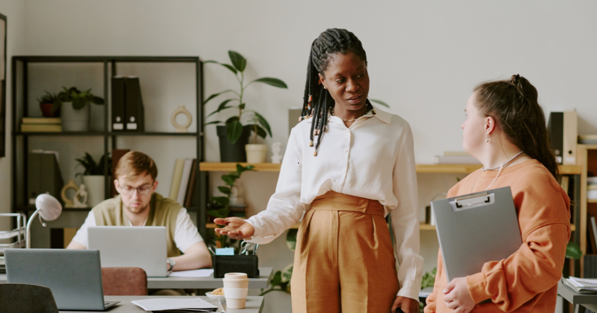 Woman in a white blouse explains something to a coworker holding a clipboard in a bright open office; two others work on laptops in the background.