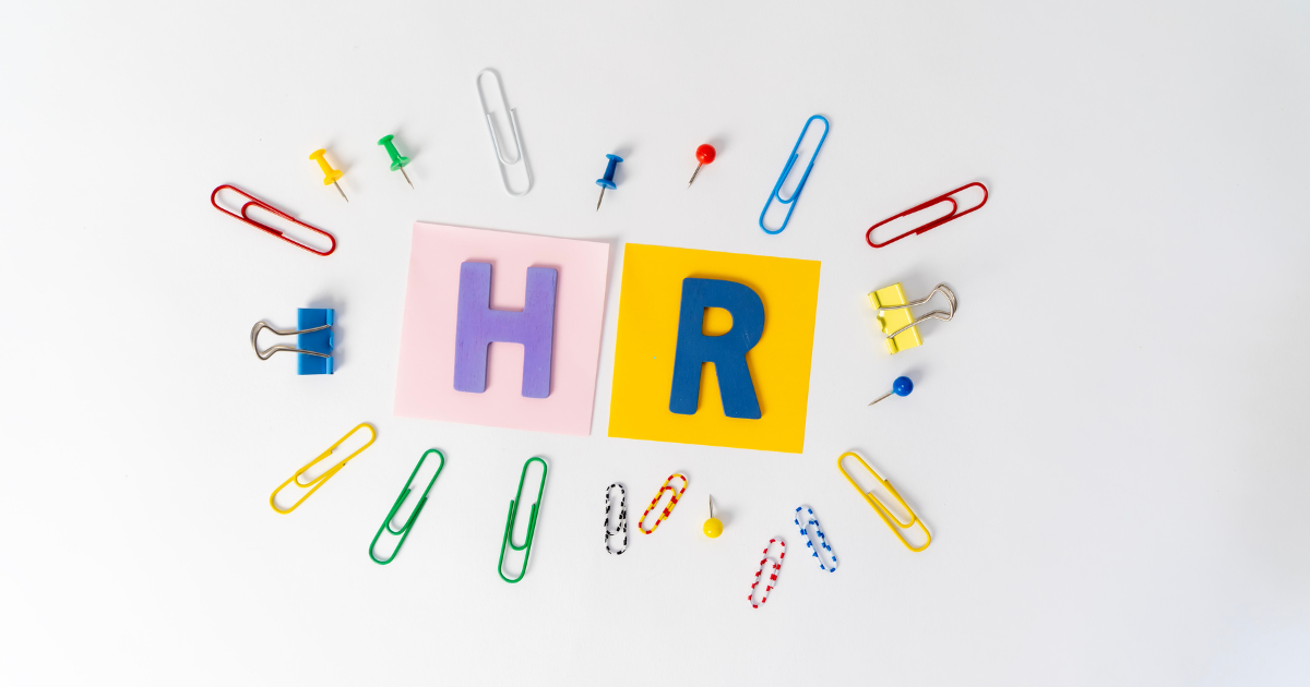 Flat lay of HR theme: pastel blocks with letters H and R surrounded by colorful binder clips, pins, and paperclips on a white background