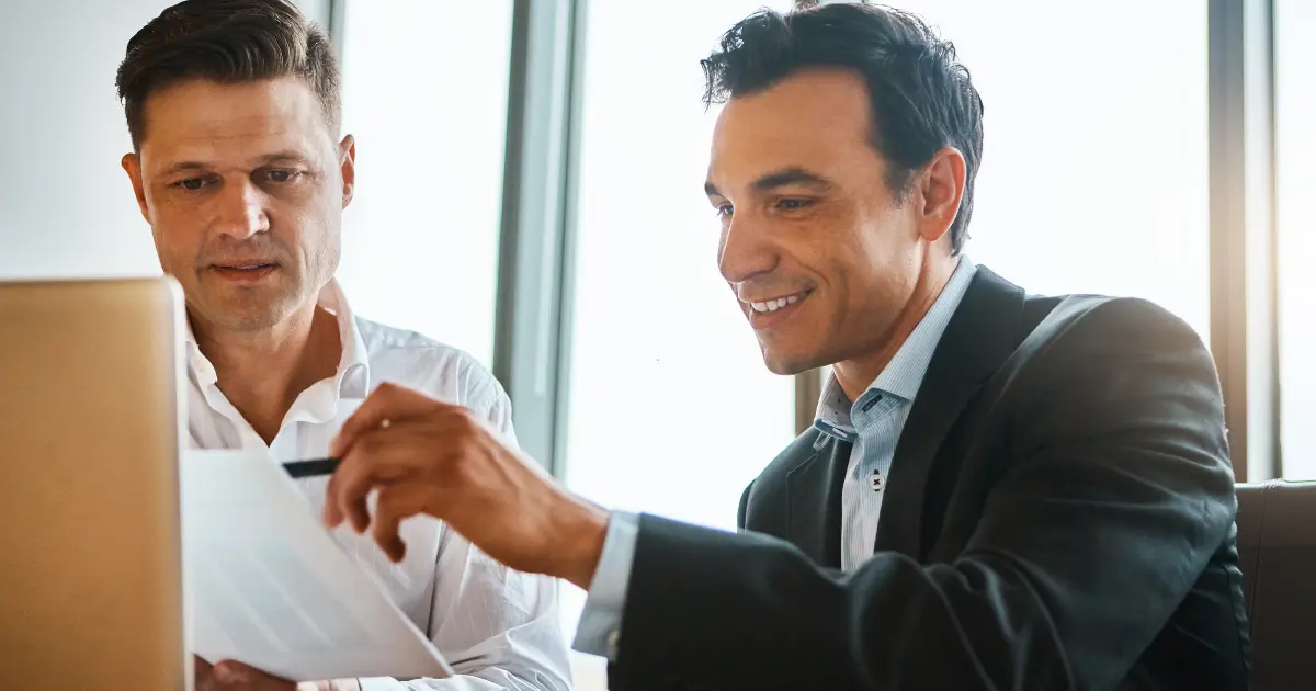 Two businessmen in a bright office review a document together beside a laptop, one pointing with a pen.