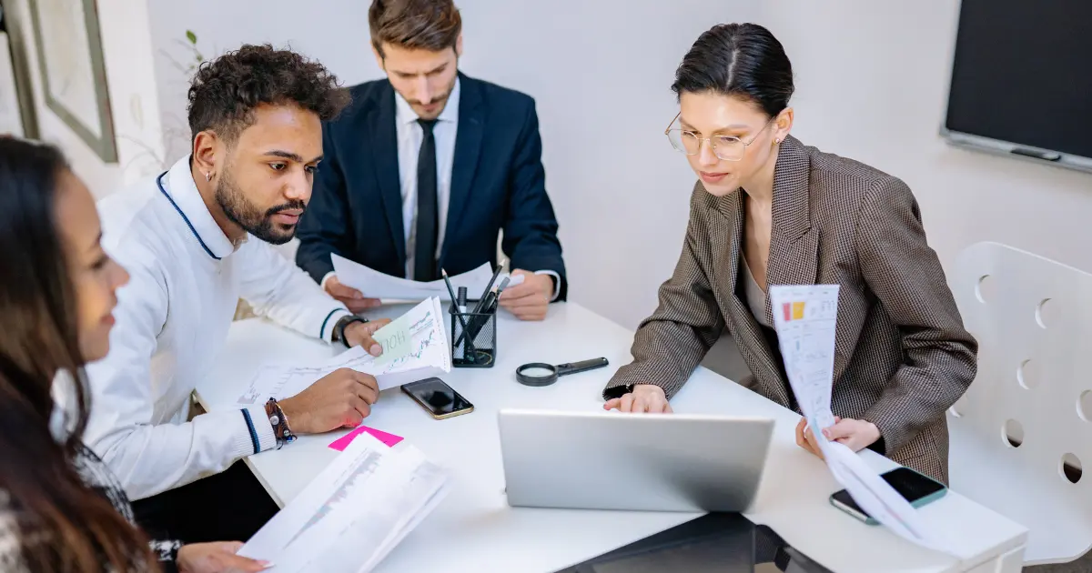 Diverse team around a white table reviewing documents; a woman in a blazer uses a laptop while others look at papers.